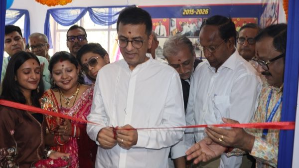 Man in a white shirt cuts a red ribbon at a ribbon-cutting ceremony, surrounded by smiling colleagues and guests in a festive setting.