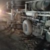 Overturned semi-truck on its side at night, with exposed wheels and trailer underneath, on a dark road surface.