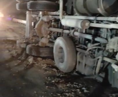 Overturned semi-truck on its side at night, with exposed wheels and trailer underneath, on a dark road surface.
