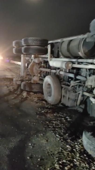Overturned semi-truck on its side at night, with exposed wheels and trailer underneath, on a dark road surface.