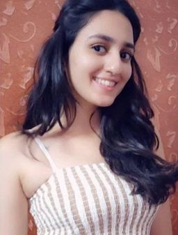 Young woman with long dark hair smiling at the camera, wearing a striped sleeveless top, in front of an orange patterned wall.