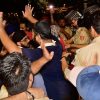 Crowd of men jostling with police officers at night, a man raises his hands while others push forward as someone films with a smartphone.