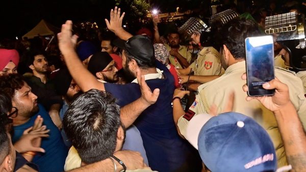 Crowd of men jostling with police officers at night, a man raises his hands while others push forward as someone films with a smartphone.