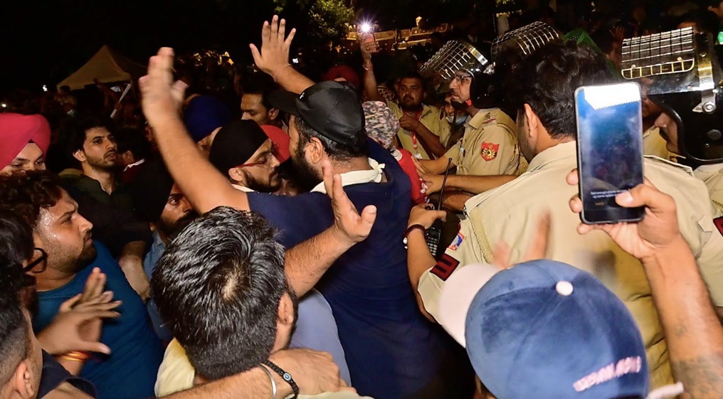 Crowd of men jostling with police officers at night, a man raises his hands while others push forward as someone films with a smartphone.