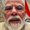 Close-up of an elderly man with a white beard and glasses taking a selfie, with an Indian flag visible in the background.