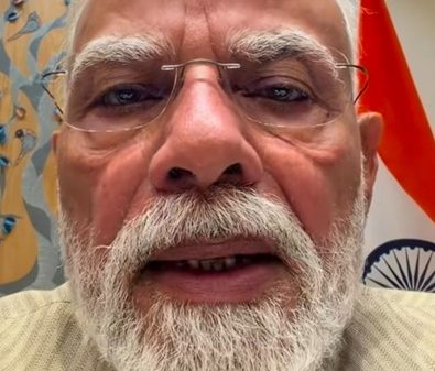 Close-up of an elderly man with a white beard and glasses taking a selfie, with an Indian flag visible in the background.