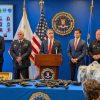 Department of Justice press conference: official at a podium with firearms laid out on a blue-covered table, flags behind.