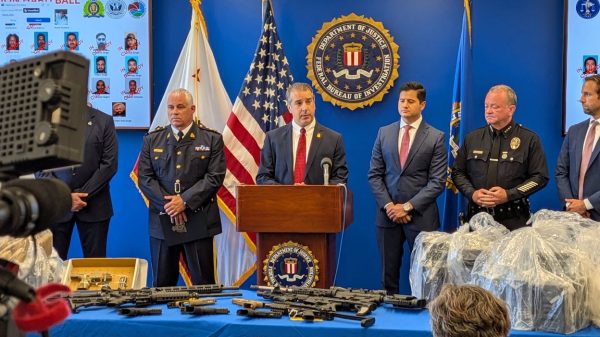 Department of Justice press conference: official at a podium with firearms laid out on a blue-covered table, flags behind.