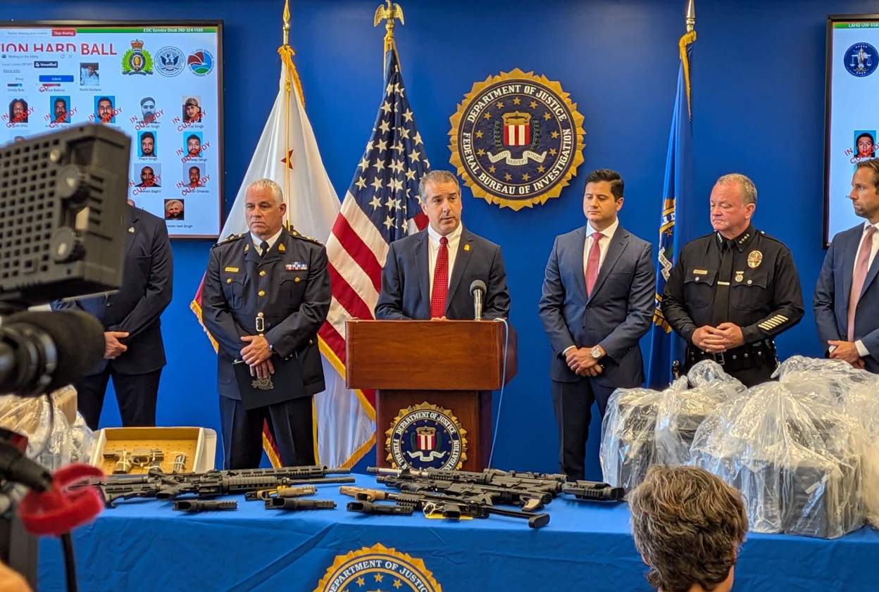 Department of Justice press conference: official at a podium with firearms laid out on a blue-covered table, flags behind.