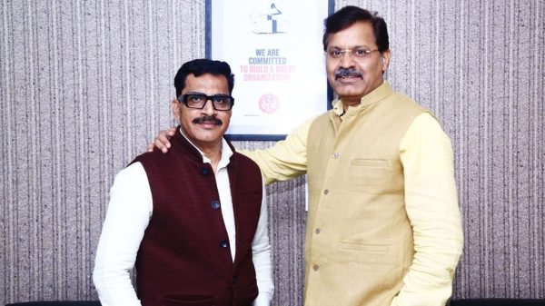 Two men pose for a photo in an office, one with his hand on the other’s shoulder; framed poster on the wall behind reads 'We are committed to build a great organization'.