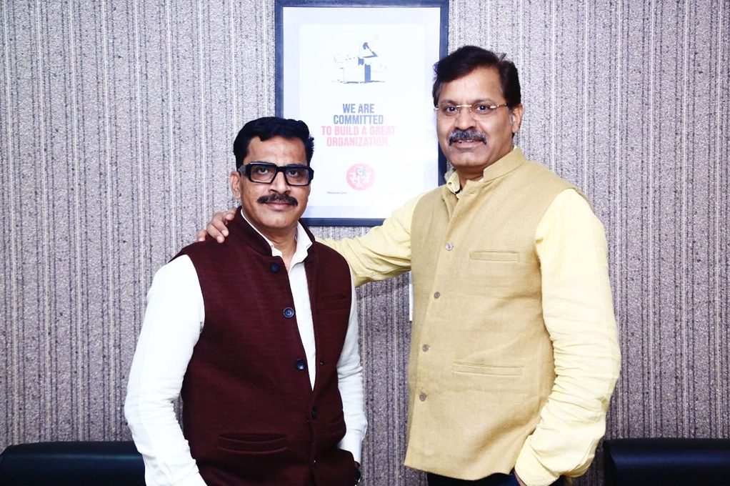 Two men pose for a photo in an office, one with his hand on the other’s shoulder; framed poster on the wall behind reads 'We are committed to build a great organization'.