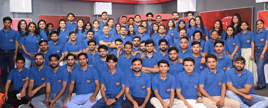 Large group of coworkers in matching blue polo shirts posing for a team photo in a studio setting with red decor in the background.