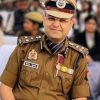 Indian police officer in khaki uniform with peaked cap, medals, and ribbons at a public event, crowd in background.