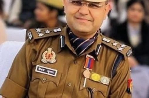 Indian police officer in khaki uniform with peaked cap, medals, and ribbons at a public event, crowd in background.