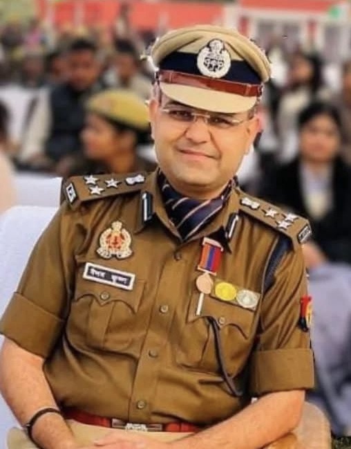 Indian police officer in khaki uniform with peaked cap, medals, and ribbons at a public event, crowd in background.
