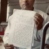 Man in a white shirt sits indoors, holding up a notebook page covered in handwritten text for display or evidence.
