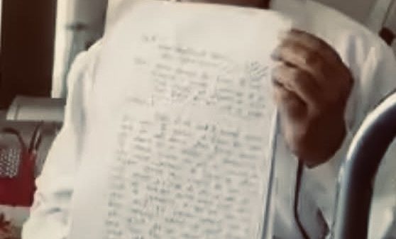 Man in a white shirt sits indoors, holding up a notebook page covered in handwritten text for display or evidence.