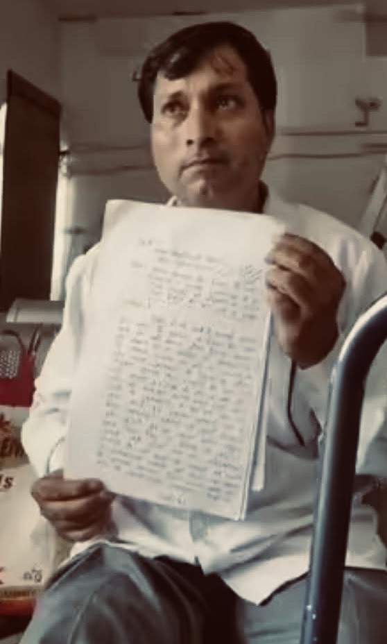 Man in a white shirt sits indoors, holding up a notebook page covered in handwritten text for display or evidence.