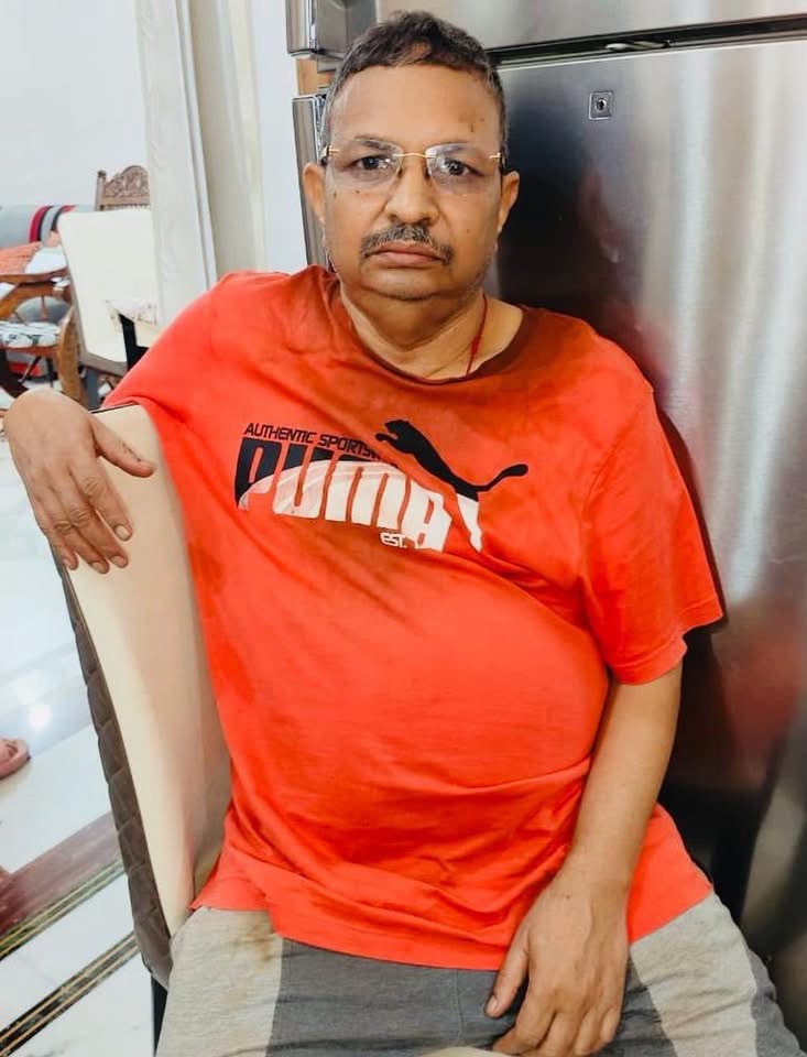 Middle-aged man with glasses and a mustache sits leaning on a chair, wearing a red Puma T-shirt in front of a stainless steel refrigerator in a kitchen area.
