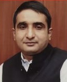 Portrait of a man with short black hair, wearing a black Nehru-style vest over a white shirt, facing the camera in a formal setting.