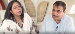 Two people seated in a private jet; a woman in a floral shawl speaks while a man with a microphone listens.