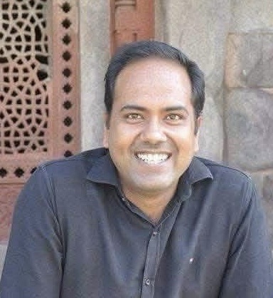 img_9682 | No. 1 Indian Media News Portal Smiling man in a dark shirt seated outdoors with a brick wall and decorative lattice behind him.