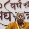 Bald man in orange robes speaking at a podium with two microphones, in front of a banner with Hindi text.