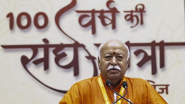 Bald man in orange robes speaking at a podium with two microphones, in front of a banner with Hindi text.
