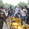 Crowd of people climbing over yellow barricades on a street; a soldier in camouflage with a rifle watches nearby; an Indian flag is visible in the crowd.