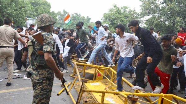 Crowd of people climbing over yellow barricades on a street; a soldier in camouflage with a rifle watches nearby; an Indian flag is visible in the crowd.