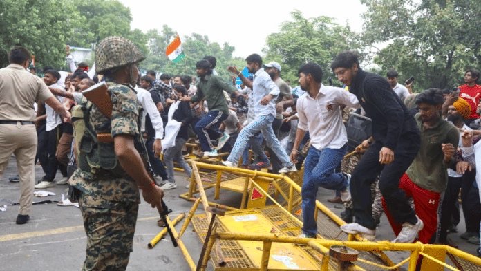 Crowd of people climbing over yellow barricades on a street; a soldier in camouflage with a rifle watches nearby; an Indian flag is visible in the crowd.