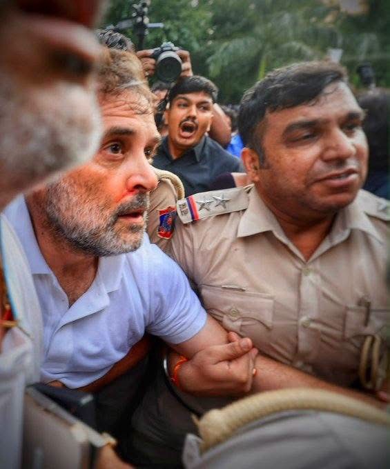 img_9867 | No. 1 Indian Media News Portal Bearded man in a light blue shirt is restrained by a uniformed officer as a photographer films the scene in a crowded outdoor area.