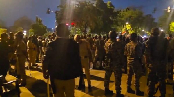 Nighttime street scene with a large crowd of civilians and uniformed officers facing each other, suggesting a protest or crowd-control event on a city street.