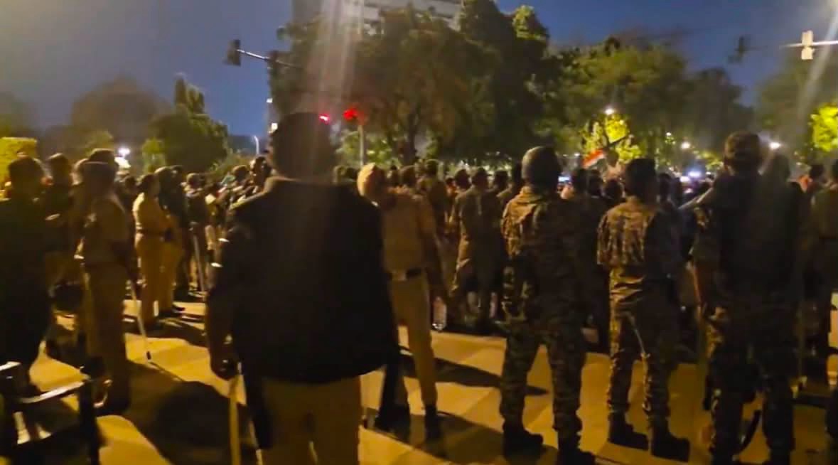 Nighttime street scene with a large crowd of civilians and uniformed officers facing each other, suggesting a protest or crowd-control event on a city street.