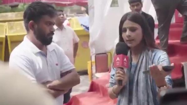 Female reporter holding a pink microphone interviewing a man with arms folded at an outdoor event.
