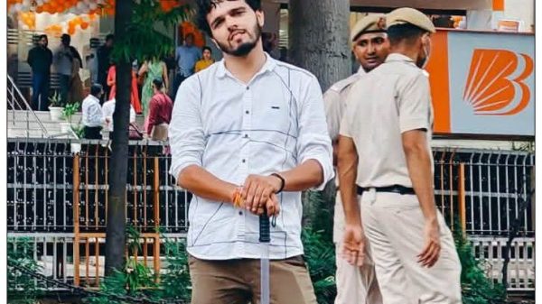 Young man in a white shirt leaning on a long cane in an outdoor market area with shoppers and security guards nearby.