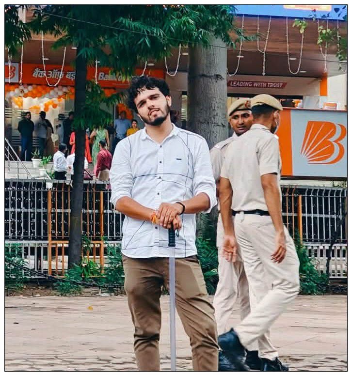 Young man in a white shirt leaning on a long cane in an outdoor market area with shoppers and security guards nearby.
