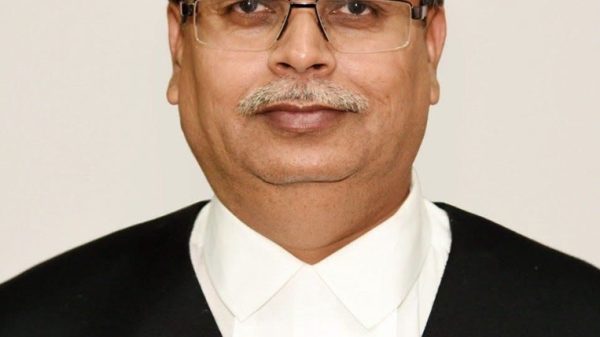 Head-and-shoulders portrait of a middle-aged man in a black suit and white shirt, wearing glasses, looking at the camera.