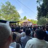 Large crowd gathered for an outdoor performance under a white-and-yellow canopy stage in a park, surrounded by trees and tents.