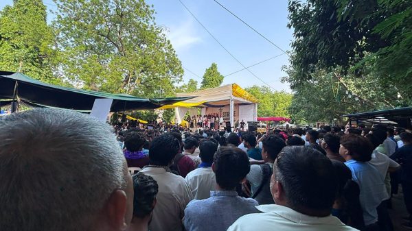 Large crowd gathered for an outdoor performance under a white-and-yellow canopy stage in a park, surrounded by trees and tents.