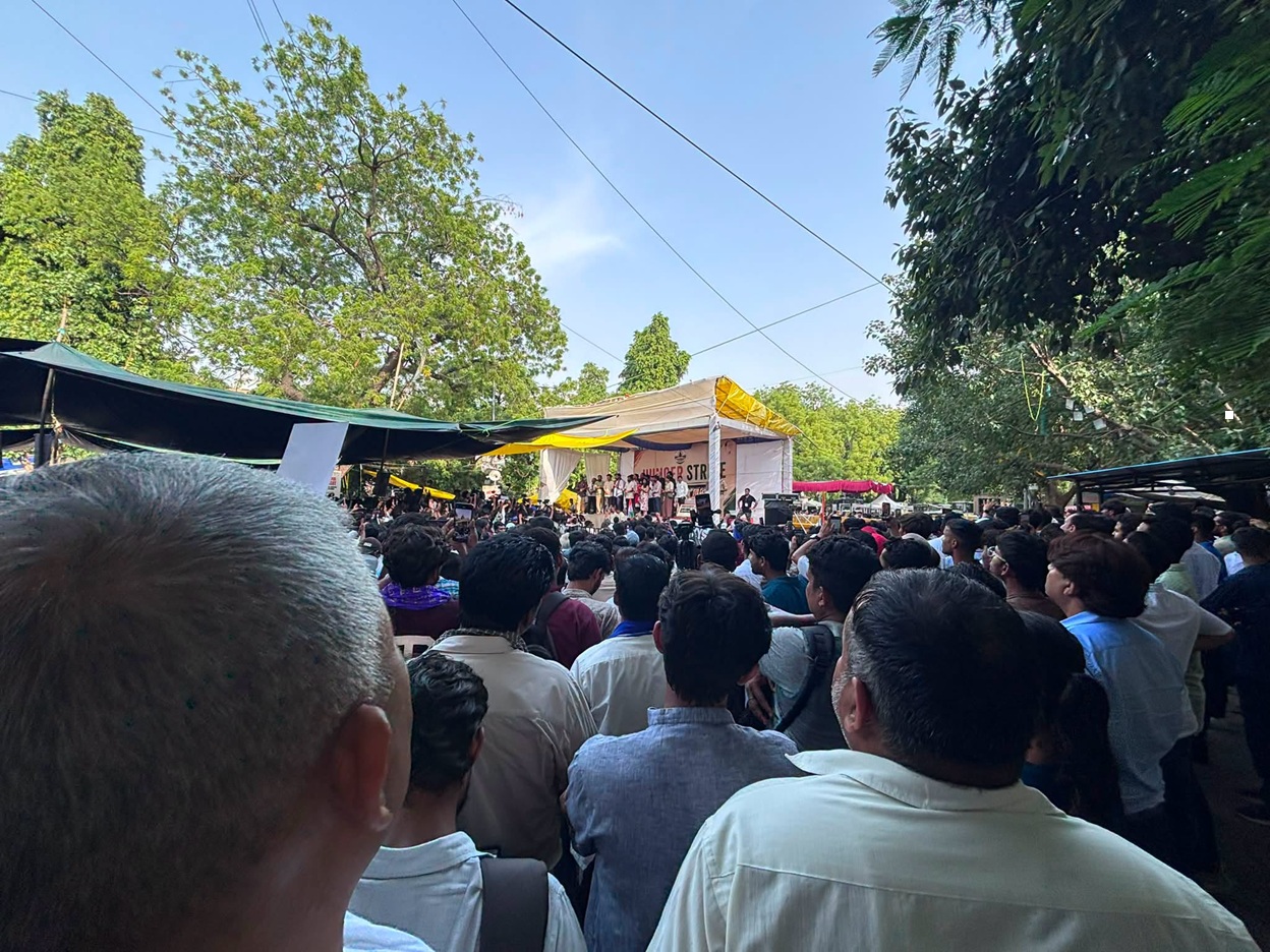 Large crowd gathered for an outdoor performance under a white-and-yellow canopy stage in a park, surrounded by trees and tents.