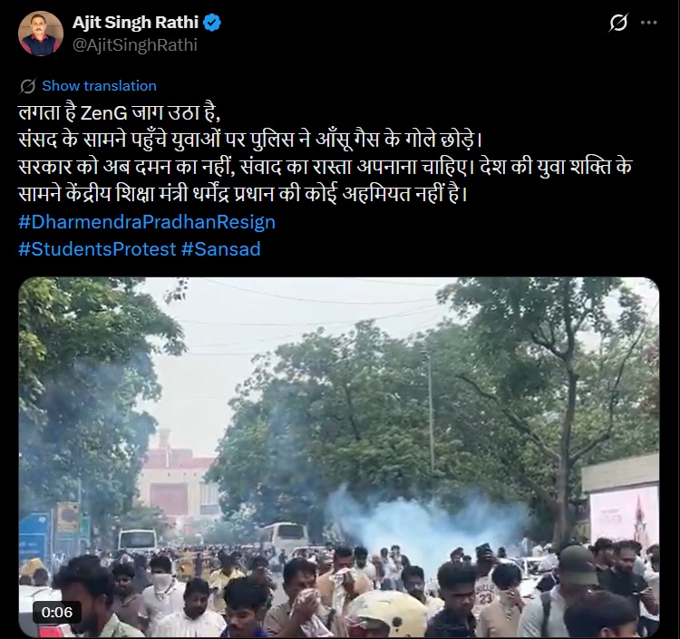 Large crowd of students protesting on a street with smoke in the air and trees lining the road, vehicles visible in the background.
