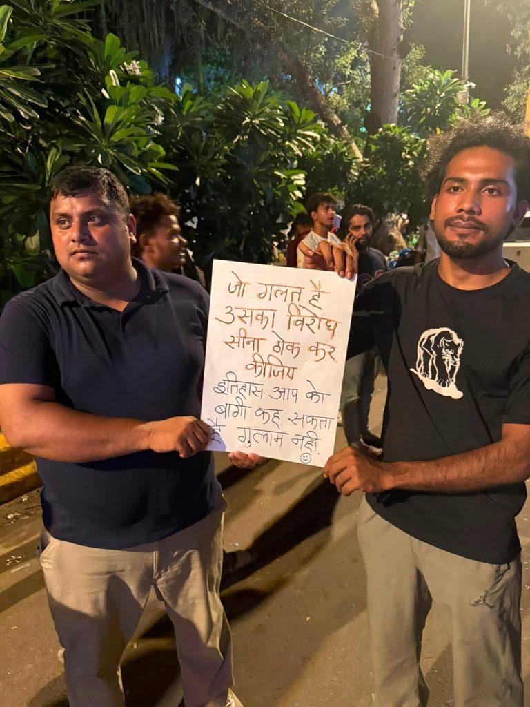 jantar mantar_1 | No. 1 Indian Media News Portal Two men on a nighttime street hold a handwritten protest sign while others stand in the background amid greenery.