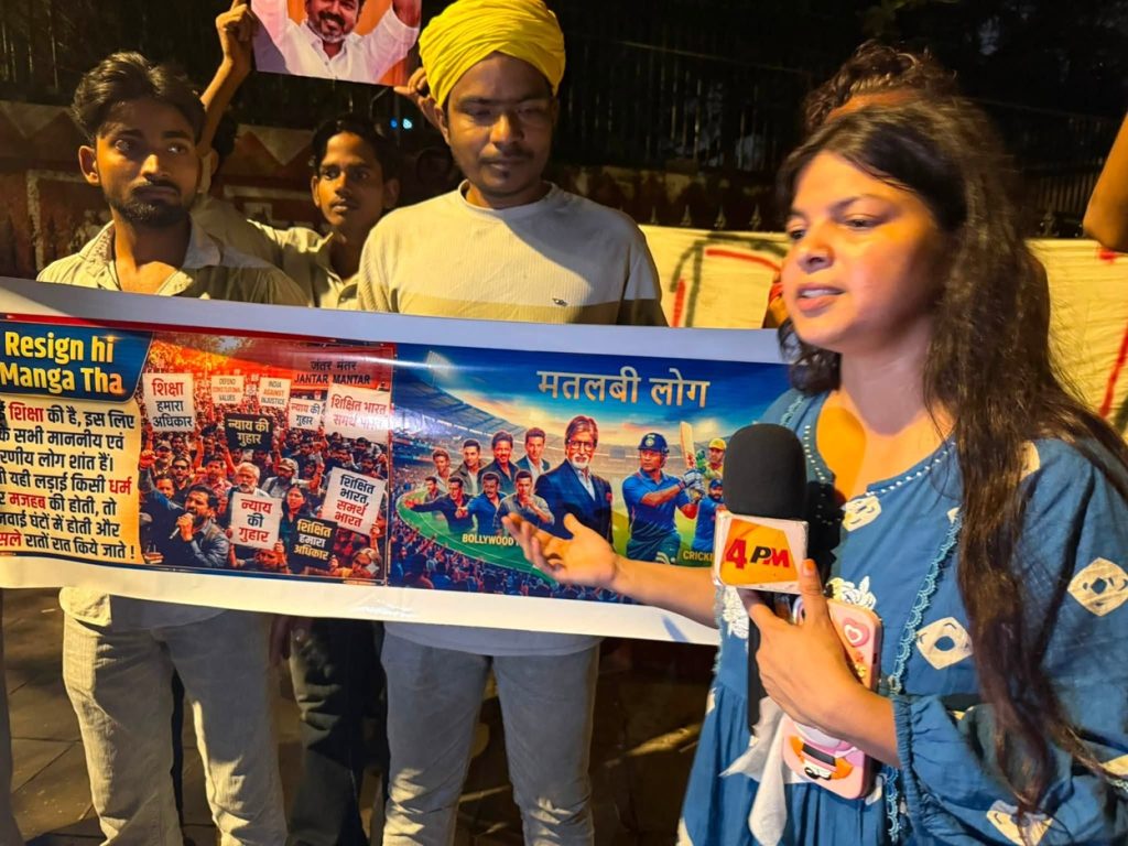 jantar mantar_2 | No. 1 Indian Media News Portal Female reporter holding a microphone speaks to protesters; a long colorful banner with Hindi text and images is held behind her at night, scene outdoors.