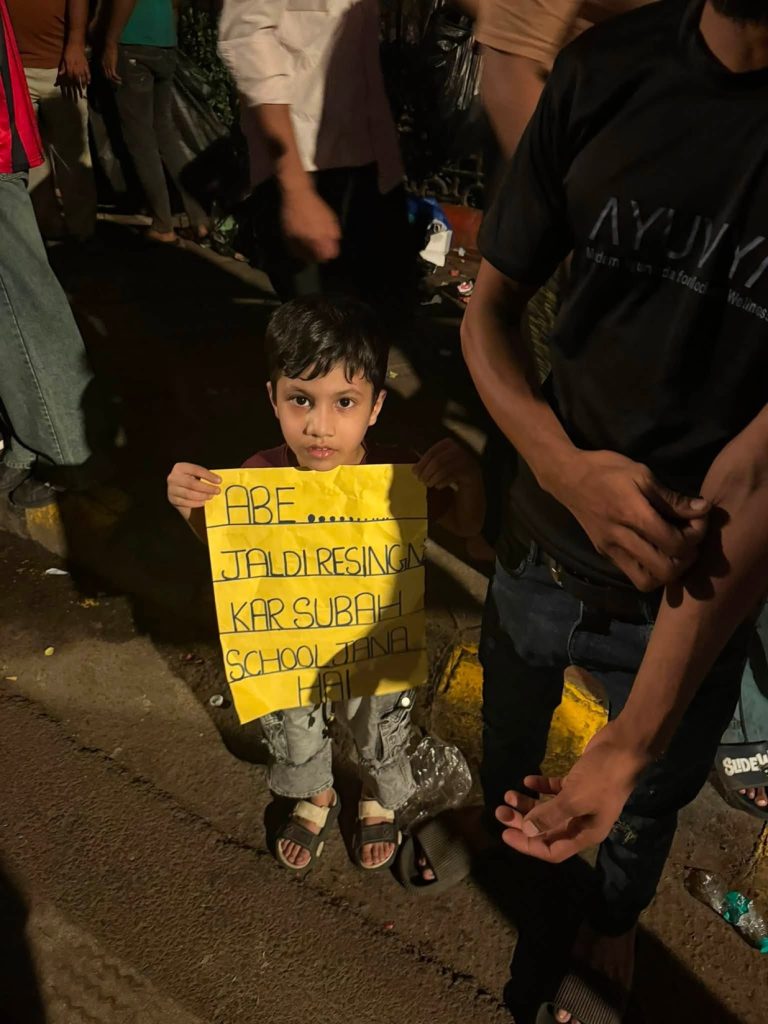 jantar mantar_3 | No. 1 Indian Media News Portal A young boy on a dimly lit street holds a yellow handwritten sign, with adults nearby in a nighttime scene.