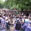Large crowd of protesters on a tree-lined street, some waving flags, confronted by riot police in blue gear.