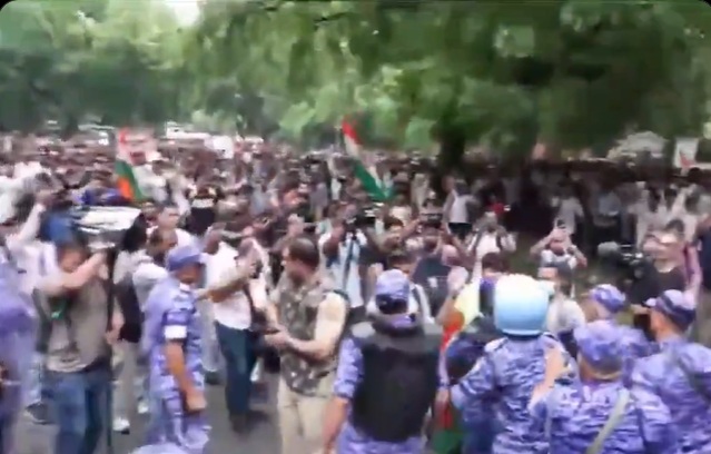 Large crowd of protesters on a tree-lined street, some waving flags, confronted by riot police in blue gear.