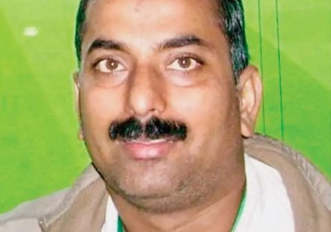 Smiling middle-aged man with short gray hair and a mustache, wearing a beige jacket and white shirt against a green background.