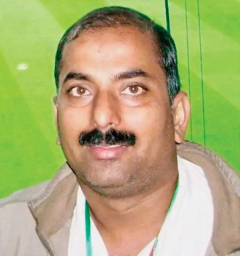 Smiling middle-aged man with short gray hair and a mustache, wearing a beige jacket and white shirt against a green background.