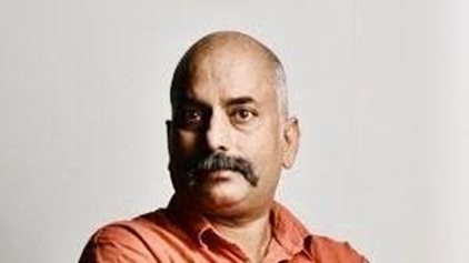 Bald man with a mustache wearing an orange shirt, looking at the camera against a light background.